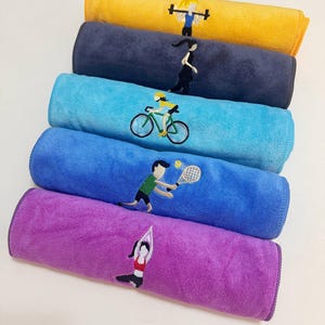 Puede incluir: Toallas enrolladas de colores con figuras de fitness bordadas. Las toallas son amarillas, azul marino, azul claro, azul oscuro y morado. Cada toalla presenta una figura bordada diferente: levantador de pesas, corredor, ciclista, tenista y practicante de yoga.