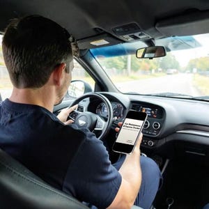 May include: A person driving a car, holding a smartphone displaying the text "Car Maintenance & Expense Tracker". The car's interior is black, with a steering wheel and dashboard visible. The road ahead is blurred.