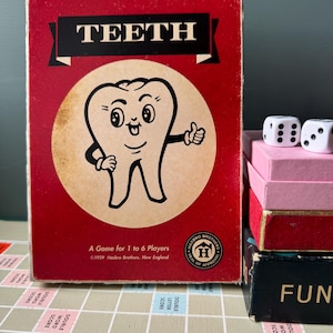May include: A vintage red board game box titled TEETH featuring a cartoon tooth character in a beige circle. The box text reads A Game for 1 to 6 Players and 1959 Hasbro Brothers, New England. Two white dice sit on stacked boxes nearby.