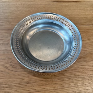 May include: A small, round, silver-colored dish with a decorative pierced border. The dish has a shallow, concave center and sits on a wooden surface. The rim features a repeating pattern of small, diamond-shaped cutouts.