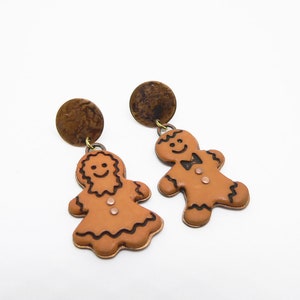 Könnte beinhalten: Ein Paar Ohrringe in Form von Lebkuchenmännern. Die Ohrringe sind aus braunem Ton gefertigt und haben eine braune Metallrückseite. Die Lebkuchenmänner lächeln und haben schwarze Umrisse.