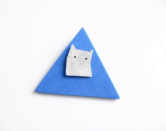 Könnte beinhalten: Eine weiße Keramik-Katzenfigur mit schwarzen Augen, die auf einem blauen dreieckigen Stück Schaumstoff sitzt.