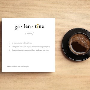 Puede incluir: Una tarjeta blanca con la palabra "galentine" en letras negras y doradas, con una definición debajo. Una taza y un platillo de café negros están sobre una superficie de madera. La definición incluye las palabras "alma gemela" y "vino".