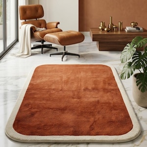 Puede incluir: Una alfombra rectangular con esquinas redondeadas, con un centro naranja quemado y un borde color crema. La alfombra está colocada sobre un suelo de mármol, con una silla y un otomán de cuero en el fondo. La alfombra añade calidez y textura a la habitación.