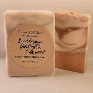 May include: Two bars of handcrafted artisan soap. One bar has a round label that reads "Sweet Orange, Patchouli & Cedarwood." The label also states "Tallow & Silk Soaps" and "Net wt. 4 oz (113.4g)." The other bar has a marbled appearance with shades of orange and cream.