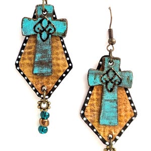 May include: A pair of handcrafted earrings featuring a turquoise cross design with a fleur-de-lis accent. The earrings have a diamond-shaped backing with a gold-colored center and black border with white dots. Dangling beads add a decorative touch.