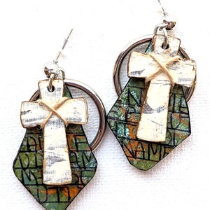 Puede incluir: Pendientes hechos a mano con un diseño de cruz blanca sobre un colgante en forma de diamante verde y negro. La cruz está atada con hilo y cuelga de un aro plateado. Los pendientes tienen un aspecto rústico y envejecido.