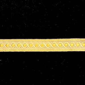 May include: A close-up of a gold-colored decorative trim with a repeating diamond pattern. The trim is set against a black background, highlighting the intricate design and texture of the woven material. This trim is suitable for various craft and sewing projects.