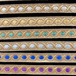 May include: Close-up of decorative trim with a repeating pattern of gold thread and colorful accents. The trim features black, white, yellow, teal, blue, purple, and pink details. The trim is likely used for embellishing clothing or craft projects.