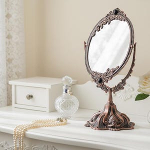 May include: A dressing table featuring an oval mirror with an ornate bronze frame, a pearl necklace, a perfume bottle, a white rose in a vase, and two small white drawers. The table is white.