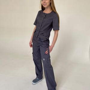 May include: A dark gray short-sleeved button-down top with a round neckline and a small pocket, paired with matching dark gray cargo pants featuring side pockets, metal D-ring accents, and adjustable drawstring hems, worn with gray clogs.