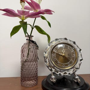 Peut inclure: Une composition décorative avec une horloge en cristal clair, un vase en verre violet et des lys roses. L'horloge a un cadran doré et repose sur une base noire. Le vase présente un motif texturé.