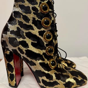 May include: Ankle boots with a leopard print pattern in black and gold. The boots have a lace-up design and are adorned with large, decorative gold buttons with black centers. The heels are high and red.