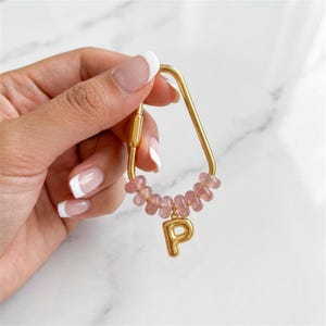 Könnte beinhalten: Goldfarbener Schlüsselanhänger mit einer Kette aus rosa Edelsteinperlen und einem goldenen Buchstaben "P" Anhänger. Der Schlüsselanhänger hat eine einzigartige, geometrische Form und einen sicheren Verschluss. Die rosa Perlen verleihen dem Accessoire einen Hauch von Farbe und Eleganz.
