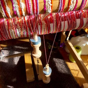 May include: A wooden weaving loom with colorful yarn in shades of red, pink, orange, and purple. The yarn is wrapped around the loom's wooden beams. Two wooden spools hang from the loom with pink thread.