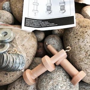 May include: A close-up of weaving loom accessories, including wooden spools with hooks, metal washers, and an instruction sheet. The sheet reads "Ashford Weaving Looms" and provides instructions for use. The items are arranged on a bed of smooth, gray stones.