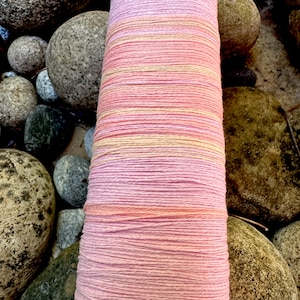 May include: A spool of pink and yellow yarn with a textured surface. The yarn is wrapped around a white cardboard core. The spool is surrounded by smooth, gray rocks.