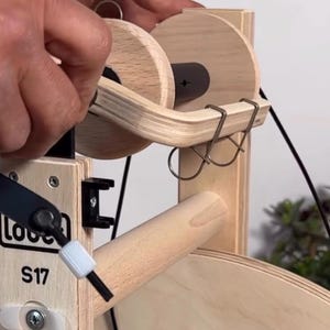 May include: A light-colored wooden spinning wheel component featuring a bobbin with two circular wooden ends, a black central core, and a curved wooden flyer arm with two metal hooks. The frame is labeled with the text Louet S17 made in holland.