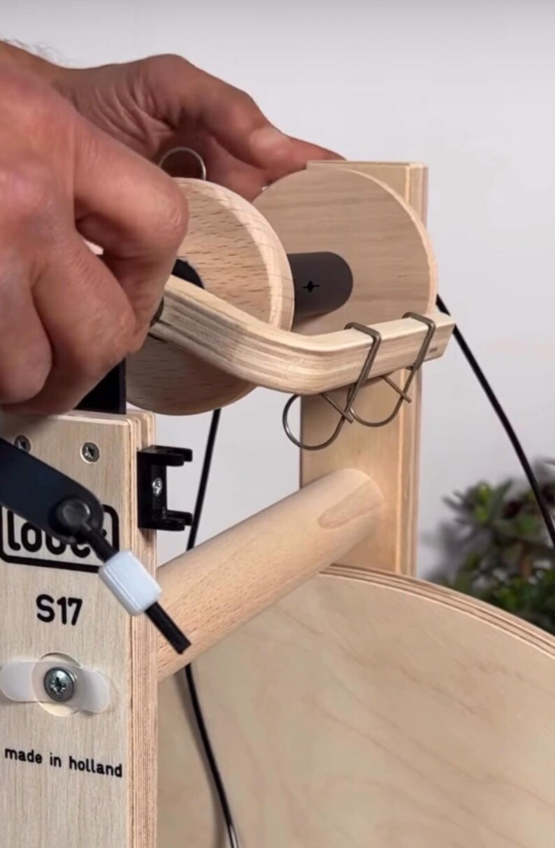 May include: A light-colored wooden spinning wheel component featuring a bobbin with two circular wooden ends, a black central core, and a curved wooden flyer arm with two metal hooks. The frame is labeled with the text Louet S17 made in holland.
