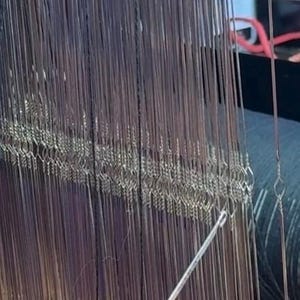 May include: Close-up of a weaving loom with numerous vertical metal heddles and warp threads. The loom is made of wood and metal, with a shuttle visible in the foreground. The image highlights the intricate process of textile creation.