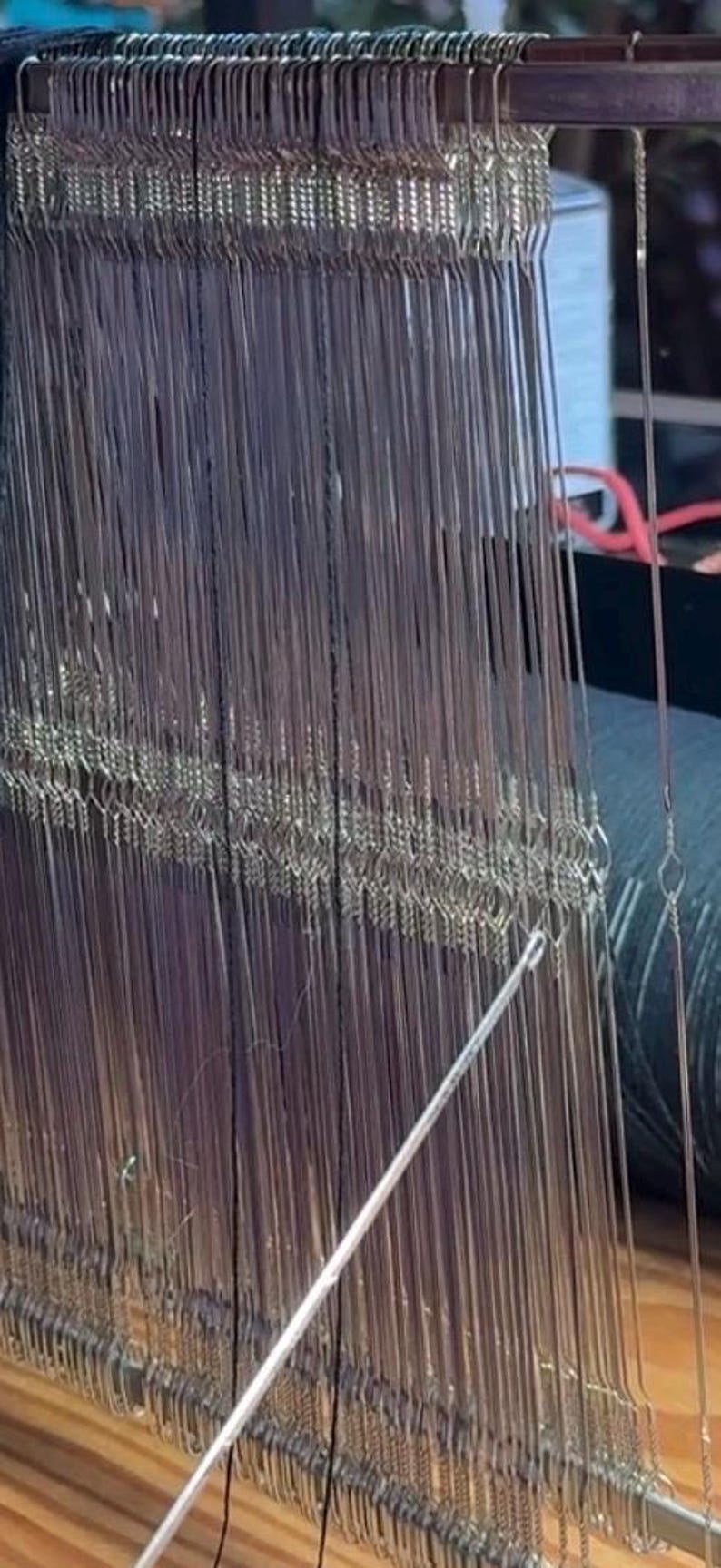 May include: Close-up of a weaving loom with numerous vertical metal heddles and warp threads. The loom is made of wood and metal, with a shuttle visible in the foreground. The image highlights the intricate process of textile creation.
