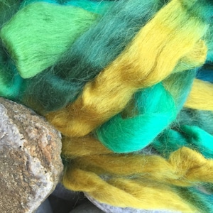 May include: Close-up of colorful fiber art in shades of green, teal, and yellow. The textured fibers are arranged in loose strands, with a few smooth stones in the foreground. The colors blend and contrast, creating a vibrant, artistic display.
