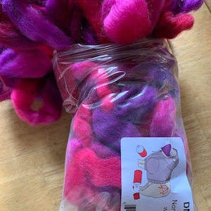 May include: A clear plastic bag filled with vibrant pink and purple wool roving. The bag has a label that reads "Northern Lights Wild Berry Jam" and "4oz". The wool is a mix of bright pink, fuchsia, and deep purple.
