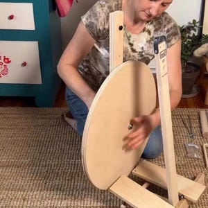 May include: A light-colored wooden spinning wheel featuring a large circular drive wheel mounted on a sturdy base with a vertical support post. The device includes a metal central hub and a vertical arm labeled with the brand name Lou&euml;t.
