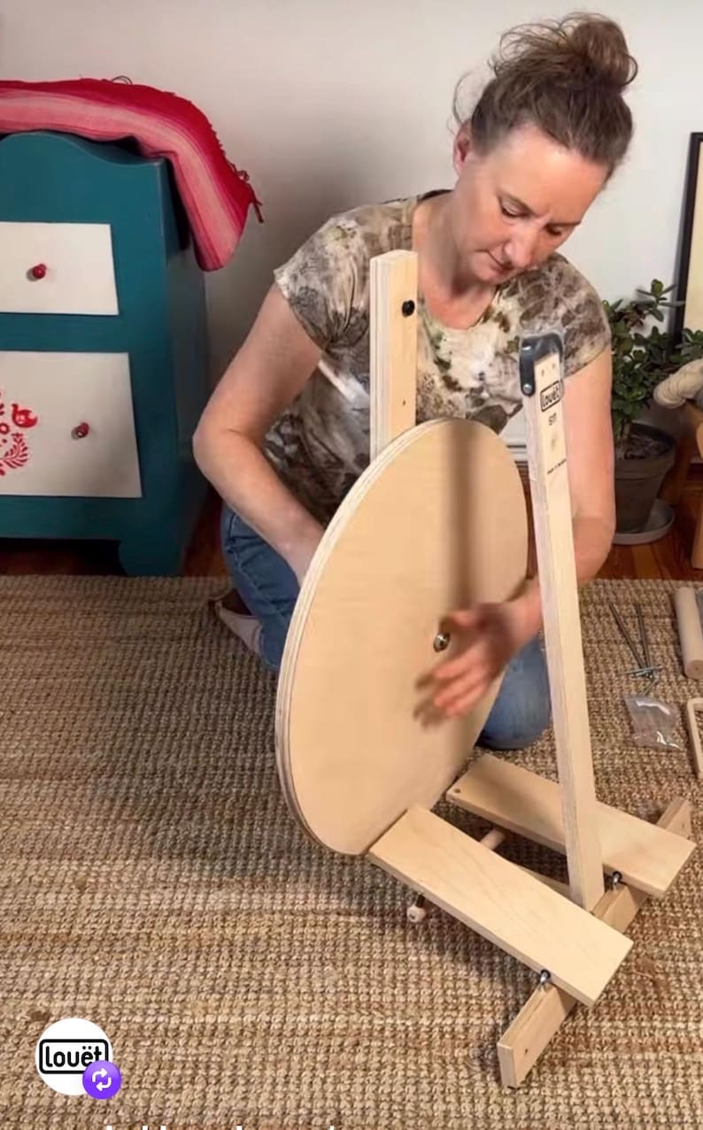 May include: A light-colored wooden spinning wheel featuring a large circular drive wheel mounted on a sturdy base with a vertical support post. The device includes a metal central hub and a vertical arm labeled with the brand name Lou&euml;t.