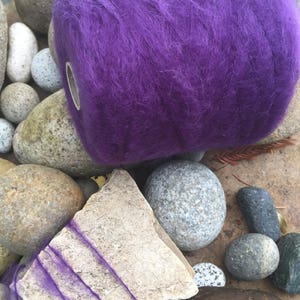 May include: A vibrant purple yarn ball with a metallic eyelet, resting among various stones. The yarn has a fluffy texture and is a deep shade of violet. A piece of stone with purple yarn wrapped around it is also visible.
