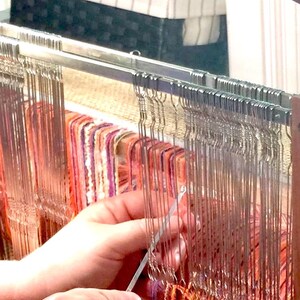 May include: Close-up of a weaving loom in use. The loom features vertical metal threads and colorful horizontal threads in shades of orange, purple, and pink. A hand is using a tool to manipulate the threads, creating a textile. A basket is in the background.
