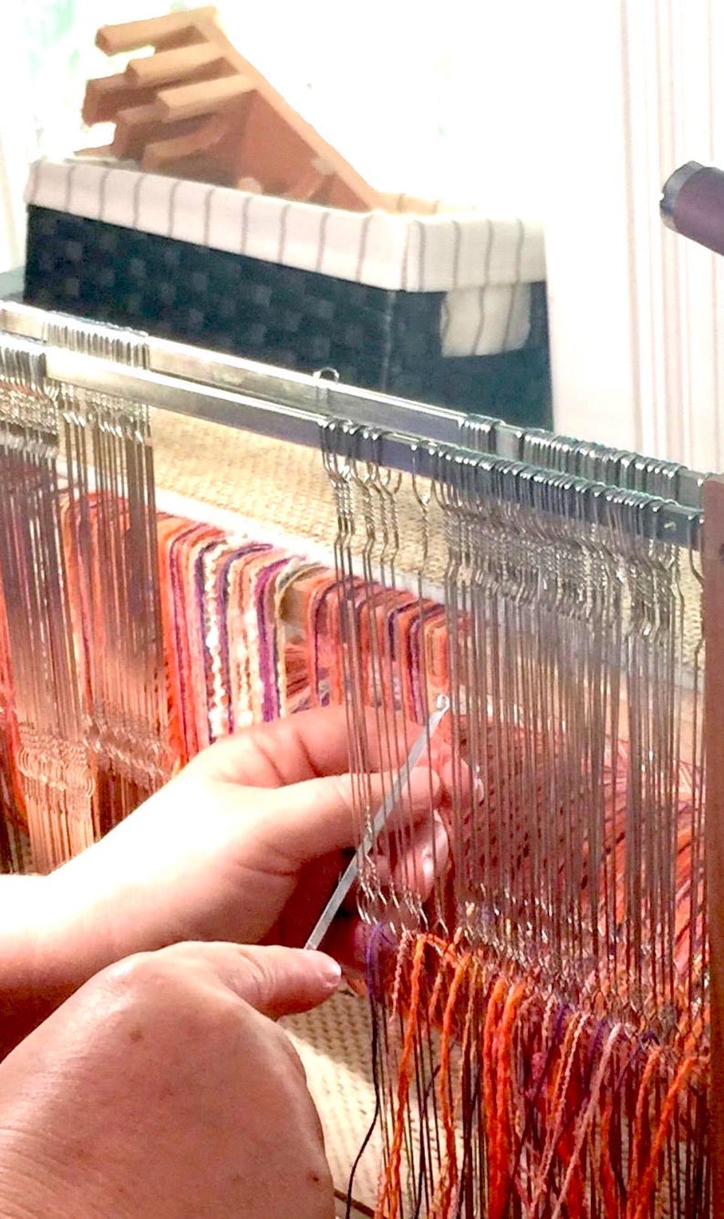May include: Close-up of a weaving loom in use. The loom features vertical metal threads and colorful horizontal threads in shades of orange, purple, and pink. A hand is using a tool to manipulate the threads, creating a textile. A basket is in the background.