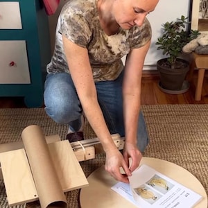 May include: A light-colored wooden circular base and a wooden assembly with a cardboard tube, accompanied by a printed instruction manual labeled S17 EN/NL/DE, featuring diagrams of a spinning wheel component on a textured beige rug.