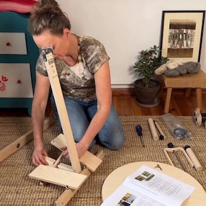 May include: A light-colored wooden Lou&euml;t spinning wheel assembly kit featuring various unfinished wood components, including a vertical support, a circular base, and a printed instruction manual, laid out on a woven floor mat for assembly.