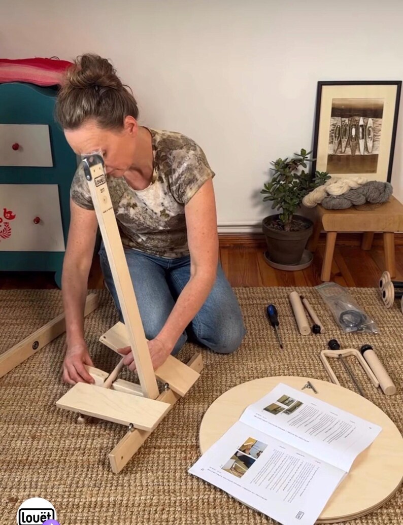 May include: A light-colored wooden Lou&euml;t spinning wheel assembly kit featuring various unfinished wood components, including a vertical support, a circular base, and a printed instruction manual, laid out on a woven floor mat for assembly.