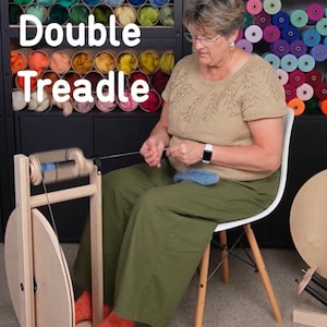 Puede incluir: Una mujer se sienta en una silla y utiliza una rueca de madera con una rueda grande de madera y una rueda m&aacute;s peque&ntilde;a con una bobina azul. La rueca est&aacute; etiquetada como "S17 Double Treadle".