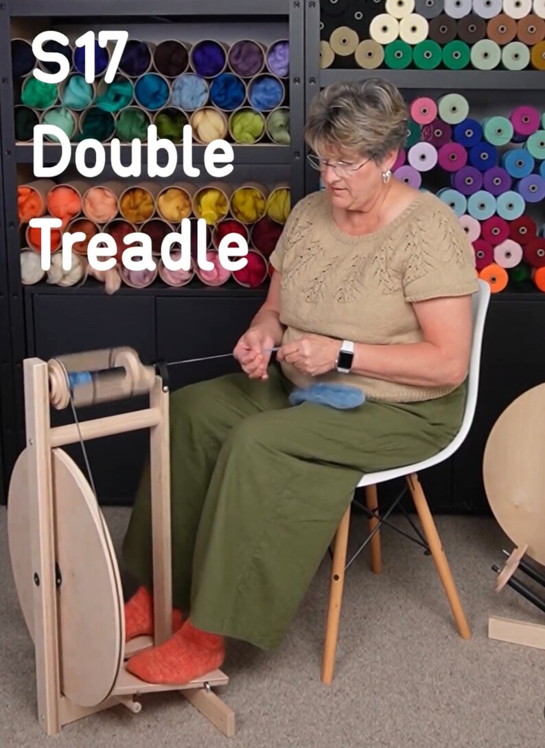 Puede incluir: Una mujer se sienta en una silla y utiliza una rueca de madera con una rueda grande de madera y una rueda m&aacute;s peque&ntilde;a con una bobina azul. La rueca est&aacute; etiquetada como "S17 Double Treadle".