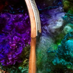 May include: A wooden handled comb with metal teeth, used for carding wool fibers. The comb is surrounded by colorful wool fibers in shades of purple, green, and blue.