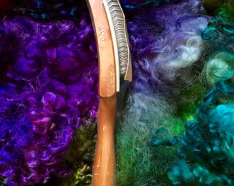 May include: A wooden fiber comb with a curved metal head, designed for processing fibers. The comb has a polished wooden handle and a series of fine metal teeth. The background features a colorful array of dyed wool fibers in shades of purple, green, and blue.