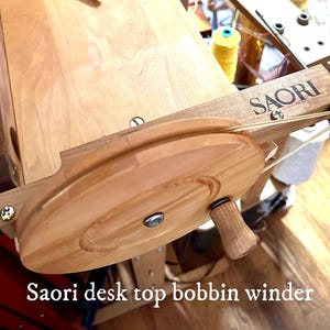 May include: A light-colored wooden Saori desk top bobbin winder featuring a circular wheel with a small handle, a metal center screw, and a thin elastic band stretched around the wheel, mounted on a wooden frame with the brand name Saori engraved.