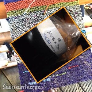 May include: A colorful woven textile in progress on a loom. The textile features a mix of purple, orange, and black threads. A roll of black thread is in the foreground, with text that reads "SAORI" and "200 x 30m".