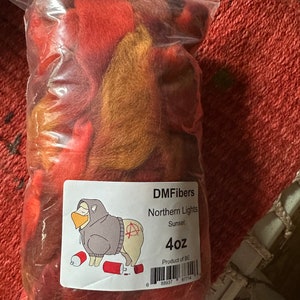 May include: A clear plastic bag filled with colorful fiber. The fiber is a blend of red, orange, and gold hues. A label on the bag reads "DMFibers Northern Lights Sunset 4oz" with a cartoon sheep wearing a hoodie and holding a spray can.