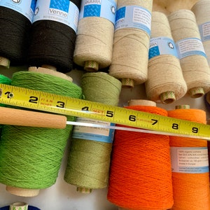 May include: A collection of yarn spools in various colors, including black, green, orange, and purple. A wooden-handled measuring tape is laid across the spools. The spools have labels that read "Venne" and "100 gr".