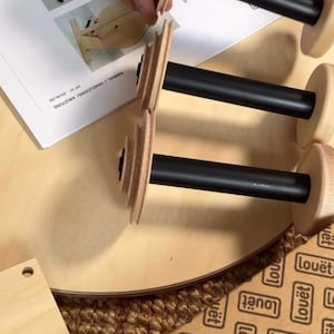 May include: Three spinning wheel bobbins with black central shafts and light-colored wooden ends, positioned near an instruction manual labeled MANUAL / HANDLEIDING / ANLEITUNG. The bobbins feature tiered wooden whorls at one end.
