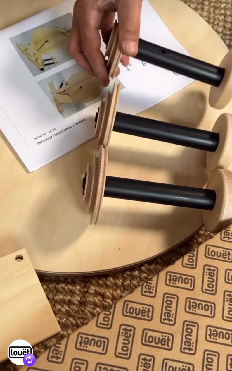 May include: Three spinning wheel bobbins with black central shafts and light-colored wooden ends, positioned near an instruction manual labeled MANUAL / HANDLEIDING / ANLEITUNG. The bobbins feature tiered wooden whorls at one end.