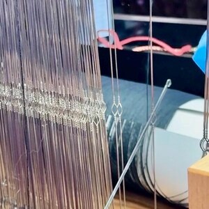 May include: Close-up of a weaving loom with numerous vertical silver threads. The threads are tightly strung, creating a dense, textured surface. A metal rod is positioned diagonally across the threads, and a wooden component is visible at the bottom.