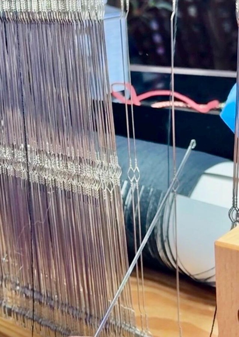 May include: Close-up of a weaving loom with numerous vertical silver threads. The threads are tightly strung, creating a dense, textured surface. A metal rod is positioned diagonally across the threads, and a wooden component is visible at the bottom.