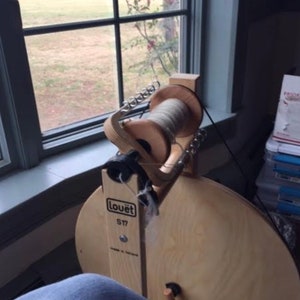 Puede incluir: Un torno de hilar Lou&euml;t S17 con un marco de madera y una gran rueda de madera. El torno est&aacute; sobre un suelo de madera y hay una ventana en el fondo.