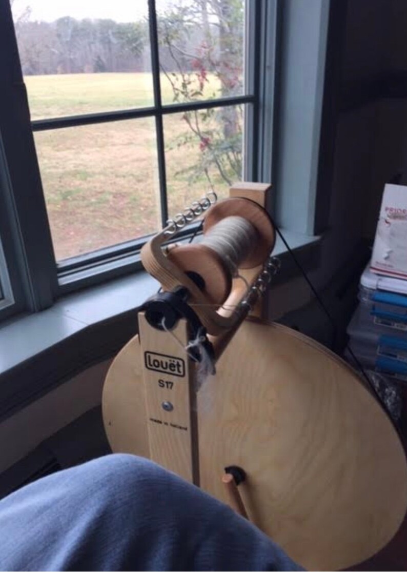 Puede incluir: Un torno de hilar Lou&euml;t S17 con un marco de madera y una gran rueda de madera. El torno est&aacute; sobre un suelo de madera y hay una ventana en el fondo.