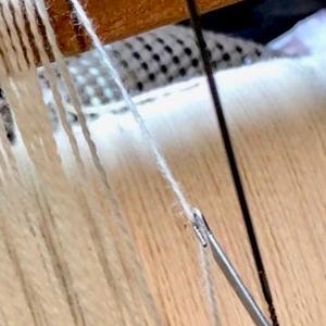 May include: Close-up of a weaving project in progress. The image shows white yarn threads being woven on a wooden loom. A metal needle is used to manipulate the threads, creating a textured fabric.
